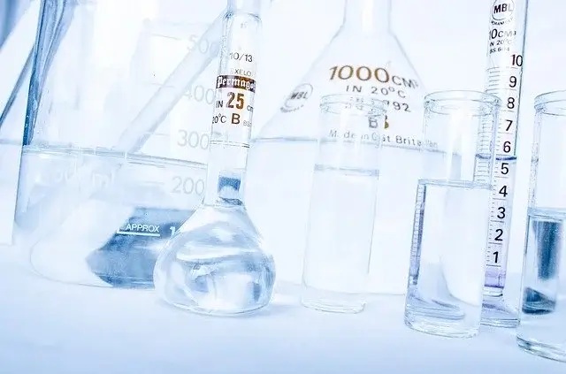 Zdjęcie przedstawia różne rodzaje szkła laboratoryjnego, takie jak kolby, cylindry miarowe i zlewki. W niektórych naczyniach znajdują się przezroczyste ciecze, a część z nich ma oznaczenia pomiarowe i etykiety, np. „1000 CM³ IN 20°C”, „25 CM³” oraz „Made in Great Britain”.